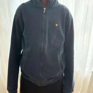 Mörkblå hoodie med dragkedja från Lyle & Scott - Säljer en mörkblå hoodie från Lyle & Scott med dragkedja framtill och klassisk gul logga på bröstet. Tröjan har huva och två fickor på sidorna. Perfekt för en avslappnad stil. Inte använd.