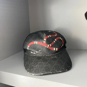Äkta Gucci keps - Snygg svart keps från Gucci med ett iögonfallande rött och vitt ormtryck på framsidan. Kepsen har diskret mönster i tyget och nät på insidan för ventilation. Perfekt accessoar för att sticka ut i mängden.