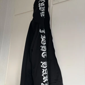 Svart zip tröja som går hela vägen (helzip) - Svart hoodie med dragkedja och vit text i gotisk stil längs båda ärmarna och huvan. Jackan har två fickor framtill och en klassisk passform. Perfekt för dig som gillar streetwear och vill sticka ut.