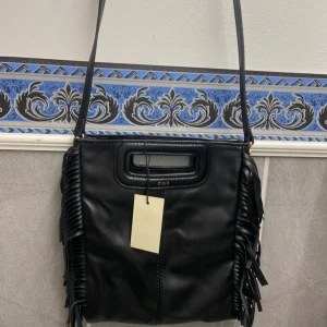  Maje väska  - Säljer en svart axelväska från Maje i skinnimitation med coola fransar på sidorna. Väskan har ett handtag upptill och en lång axelrem, samt diskret guldfärgad logga.  Helt ny.  Dustbag medföljer.