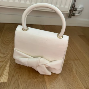 Vit handväska med rosett - Säljer en stilren vit handväska med ett stort handtag och en dekorativ rosett framtill. Väskan har en klassisk, fyrkantig form och passar perfekt för att lyfta din outfit.