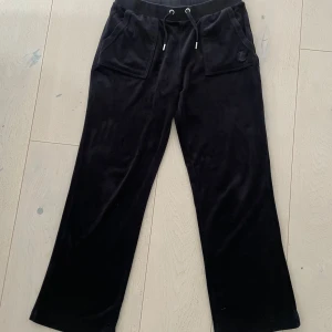 Svarta mjukisbyxor från Juicy Couture - Säljer ett par svarta mjukisbyxor från Juicy Couture med breda ben, dragsko i midjan och stora fickor fram och bak. Klassisk logga på ena fickan framtill. Byxorna är upplagda en gång och har innerbenslängden 76 cm. Perfekta för en avslappnad och trendig look. Byxorna är i storlek 15/16 år med passar dig som har storlek S. 