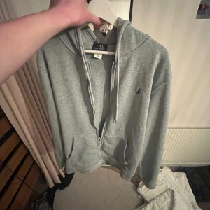 Grå hoodie  - Säljer en klassisk grå hoodie från Polo  med dragkedja och huva. Tröjan har två fickor framtill och en liten broderad logga på bröstet. Perfekt för en avslappnad stil.