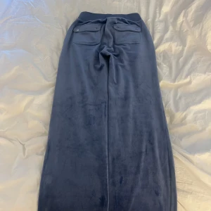 Grå blåa Juciy Couture byxor  - Mörkblå mjukisbyxor i mjukt material med bred resår i midjan och dragsko. Byxorna har raka, vida ben och två fickor med lock och knapp både fram och bak. Perfekta för en avslappnad stil.