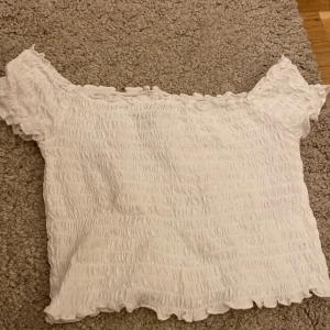 Vit smockad offshoulder topp - Säljer en söt vit offshoulder topp med smockat tyg och korta ärmar. Toppen har en croppad passform och vågiga kanter upptill och nedtill. Perfekt att styla till jeans eller kjol!