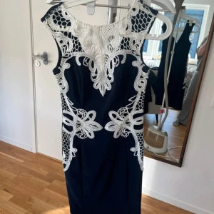 Mörkblå klänning med vit broderad spets - Säljer en mörkblå klänning med fantastisk vit broderad spetsdetalj upptill och längs sidorna. Ärmlös modell med rund halsringning och figurnära passform. Perfekt för speciella tillfällen!