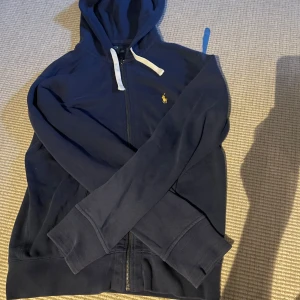  Ralph Lauren Zip Hoodie - Säljer en stilren marinblå hoodie från Polo Ralph Lauren som är perfekt för sommarkvällar i storlek S. Den är lite solblekt men inget man direkt tänker på. Skick 7/10. Nypris ligger på ca 2000kr. Pris diskuterbart vid snabb affär!!