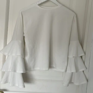 Vit långärmad topp med volangärmar - Säljer en vit långärmad topp med unika volangärmar i flera lager. Perfekt för dig som vill sticka ut med en trendig och stilren look. Toppens material känns mjukt och bekvämt mot huden.