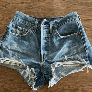 Blå jeansshorts från Levi's - Säljer ett par klassiska blå jeansshorts från Levi's med hög midja och slitna kanter. Shortsen har fem fickor och en vintagekänsla med slitningar framtill. Perfekta för sommaren!
