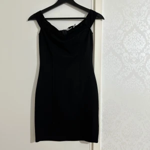 Svart offshoulder klänning från deJara x NA-KD - svart, tight offshoulder klänning från deJara x NA-KD i storlek XS. Klänningen är kort, har en smickrande passform och dragkedja bak. Perfekt för dig som gillar stilrena och eleganta plagg.