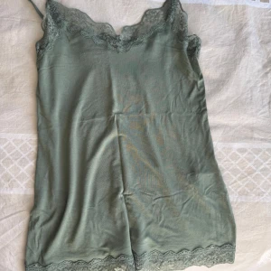 Grön linnetopp med spetsdetaljer - Snygg grön linnetopp med tunna axelband och spets längs både ringning och nederkant. Perfekt för sommardagar eller som lager under en tröja. Mjuk och lätt i materialet. Helt oanvänd💞