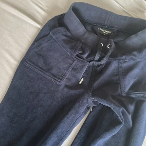 Mörkblå mjukisbyxor från Juicy Couture - Mörkblå mjukisbyxor från Juicy Couture med snörning i midjan och stora fickor med knappar bak. Byxorna har en mjuk och skön känsla, perfekta för en avslappnad stil. Klassisk logga på insidan av linningen.