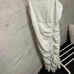 Vit långklänning med spetsdetaljer - Elegant vit långklänning med smala axelband och vackra spetsdetaljer. Klänningen har en åtsittande passform med rynkade detaljer längs sidan som ger en fin siluett. Perfekt för sommarens festligheter. Storlek L men skulle säga att den är i storlek M.