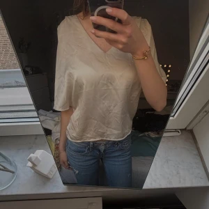 Beige v-ringad blus - Säljer en beige v-ringad blus med lös passform och korta ärmar. Perfekt till jeans eller kjol för en avslappnad stil.