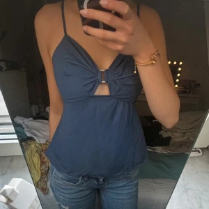 Blå linnetopp med ringdetalj - Säljer en blå linnetopp med smala axelband och snygg ringdetalj framtill. Toppen har en liten cut-out under bysten och är lätt utsvängd nertill. Perfekt för varma dagar!