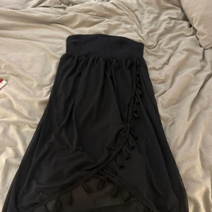 Svart strand klänning - Säljer en svart strandklänning med bred smock vid brösten och dekorativa tofsar längs kanten. Klänningen har volangdetaljer och är gjord i ett lätt, transparent tyg som ger en luftig känsla. 