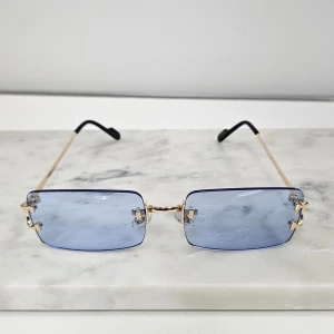 Rektangulära solglasögon med blåtonade glas - Snygga solglasögon med rektangulära, blåtonade glas och tunna guldfärgade bågar. Glasögonen har minimalistisk design utan synlig ram runt glasen och svarta detaljer vid skalmarna. Perfekt accessoar för en trendig look.