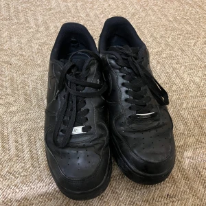 Svarta Nike Air Force 1 sneakers - Klassiska helsvarta Nike Air Force 1 sneakers med svart snörning och rund tå. Skorna har en platt sula och är tillverkade i skinn med diskret Nike-logga på sidan. Perfekta för en stilren och sportig look.