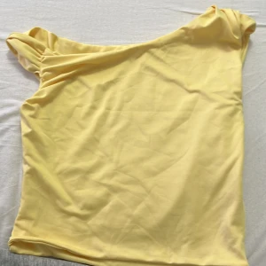 Gul oneshoulder topp - Snygg gul topp med oneshoulder-design och lätt rynkade detaljer vid axeln. Perfekt för dig som vill sticka ut med en enkel men trendig look. Tunn och mjuk känsla, passar till både jeans och kjol.