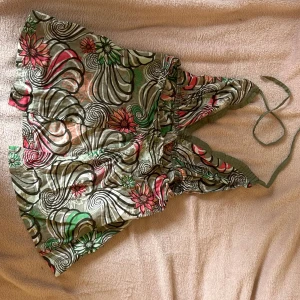 Babydoll topp med halter neck - Säljer en färgglad topp med blommigt och swirl-mönster i grönt, rosa, rött och beige. Toppen har knyt i nacken och är ärmlös med omlott-v-ringad front. Perfekt för sommarens varma dagar och har en lätt och luftig känsla. Öppen i ryggen! Resonen är lite sönder! 