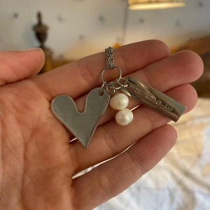 Långt silverhalsband med hjärta & pärlor - Unikt långt halsband i silverfärgad metall med tre hängen: ett stort hjärta, två vita pärlor och en rektangulär bricka med ingraverad text. Kedjan är ca 37 cm lång och ger en cool statement-look. Perfekt för dig som gillar personliga smycken med lite edge.