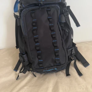 Svart och blå Integral 45 ryggsäck - Stor svart ryggsäck med blå detaljer och broderad logga från NCC Integral. Ryggsäcken har flera justerbara remmar, vadderade axelband och höftbälte för extra komfort. Perfekt för längre äventyr med smarta fästen och många fickor. Volymen är 45 liter.