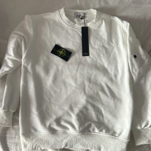 Vit sweatshirt från Stone Island - Vit sweatshirt från Stone Island med klassisk rund hals och ribbade muddar. Tröjan har den ikoniska Stone Island patchen som kan fästas på ärmen. Tillverkad i mjuk bomull, perfekt för en clean och stilren look. Passar dig som gillar streetwear och exklusiva märken.