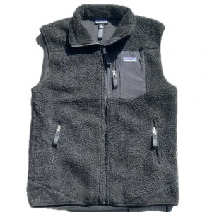 Svart fleeceväst från Patagonia - Svart fleeceväst från Patagonia med hög krage och hel dragkedja framtill. Västen har två sidofickor med dragkedja och en bröstficka med dragkedja och logga. Mjuk teddyfleece och sportig look, perfekt för lager-på-lager.
