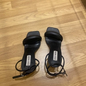 Svarta klack-sandaler Steve Madden - Säljer ett par svarta sandaler från Steve Madden med hög klack och fyrkantig tå. Skorna har en bred, vadderad rem över tårna och smala remmar runt ankeln med spänne. Tillverkade i skinn för en stilren och trendig look.