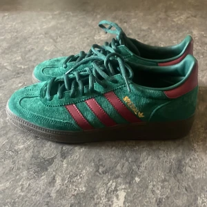 Adidas Spezial grönt och vinrött - Säljer ett par supersköna Adidas Spezial sneakers i grön mocka med vinröda läderdetaljer och klassiska tre ränder på sidan. Skorna har snörning, rund tå och platt sula. 