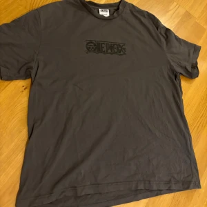 One Piece Zoro t-shirt  - Säljer en mörkgrå t-shirt med One Piece-tryck fram och stor Zoro-illustration på ryggen. Skön passform och tillverkad i mjuk bomull. Perfekt för dig som älskar anime och vill ha en unik tee med coolt motiv.