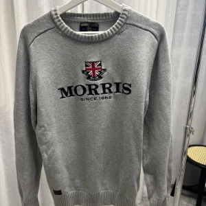 Grå stickad tröja från Morris Gents - Snygg grå stickad tröja från Morris Gents med broderad logga och brittisk flagga på bröstet. Tröjan har rund halsringning, ribbade muddar.