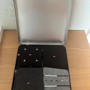 Tommy Hilfiger strumpor 4-pack presentbox 39-42 - Fyra par snygga strumpor från Tommy Hilfiger i svart, grått och ljusgrått. Enkla och stilrena med logotyp, ett par med diskret mönster och ett par med ränder och text. Kommer i en exklusiv presentbox. Material: 77% bomull, 21% polyamid, 2% elastan. Storlek EU 39-42.