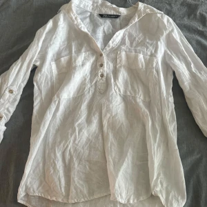 Vit linneskjorta från Zara - Vit långärmad skjorta från Zara i luftigt linne. Skjortan har två bröstfickor, knappar framtill och avslappnad passform. Perfekt för varma dagar och enkel att styla med jeans eller shorts.