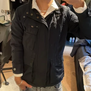 Svart parkajacka med fickor från G-STAR - Snygg svart parkajacka från G-STAR med flera praktiska fickor och dragkedja framtill. Jackan har hög krage, knappdetaljer och är långärmad. Perfekt för dig som vill ha en stilren och funktionell jacka till vardags.