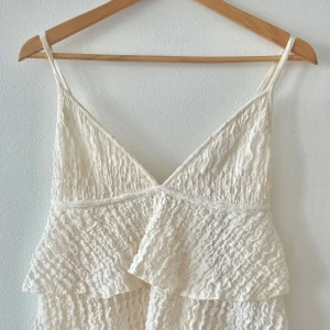 Vit volangtopp från Gina Tricot - Supersöt vitt linne från Gina Tricot med tunna axelband och v-ringning. Linnet har volangdetaljer och ett vågigt, strukturerat tyg som ger en romantisk känsla. Perfekt för varma dagar och somriga vibes.