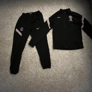 Svart PSG träningsväst från Nike - Svart träningsväst från Nike x PSG med dragkedja framtill och vita detaljer på ärmarna. Västen har PSG-logga och Nike Swoosh på bröstet. Sportig look med hög krage och smal passform, perfekt för fotbollsträning eller chill.