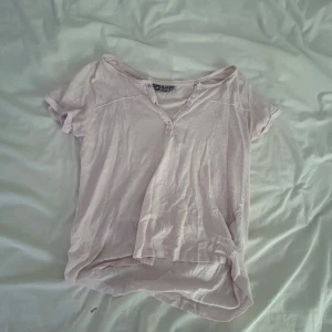 Ljusrosa t-shirt från Red & Blue - Ljusrosa t-shirt från Red & Blue med korta ärmar och knappar vid halsringningen. T-shirten har en avslappnad passform och är tillverkad i mjuk bomull, perfekt för en chill och enkel stil.