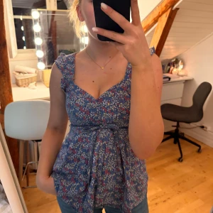 Blommig blå blus från H&M - Supersöt blommig blus från H&M i blått med små röda, vita och gula blommor. Modellen har volangdetaljer vid axlarna, v-ringning och knytband i midjan som ger en fin siluett. Perfekt för vår och sommar!