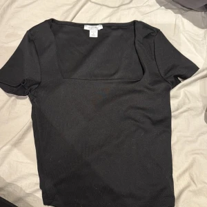 Svart topp med fyrkantig ringning Amisu - Svart kortärmad topp från Amisu med snygg fyrkantig ringning. Topp i stretchigt material som sitter skönt och har en enkel, stilren look. Perfekt att matcha med jeans eller kjol för en trendig outfit.