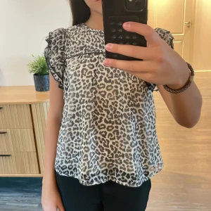 Leopardmönstrad blus från Lindex - Säljer en luftig blus från Lindex med svart och grått leopardmönster. Toppen har volangärmar med svarta spetsdetaljer och är tillverkad i ett tunt, lätt material.