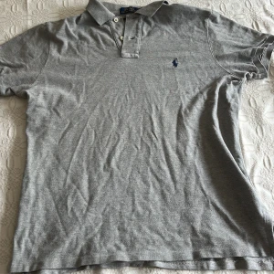 Grå pikétröja från Polo Ralph Lauren - Klassisk grå pikétröja från Polo Ralph Lauren med broderad logga på bröstet. Tröjan har krage, knappar framtill och är tillverkad i mjuk bomull. Custom fit-modell som sitter snyggt och passar till det mesta. Priset går att diskutera 🙌