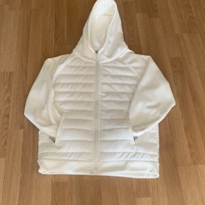 Vit quiltad pufferjacka med huva - Snygg vit pufferjacka från LAI med quiltad framsida och mjuka långärmade ärmar. Jackan har huva, hel dragkedja och två sidofickor med dragkedja. Perfekt för dig som gillar stilren och clean look. Materialet är polyester och syntet.