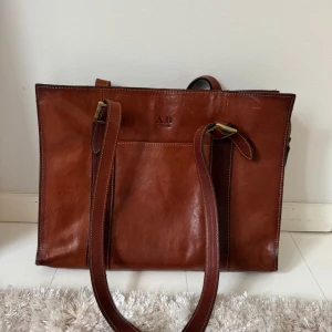 Brun axelväska i skinn från A.D Firenze - Snygg brun axelväska i skinn från A.D Firenze med stilrena detaljer och rejäla axelremmar. Väskan har flera fack med dragkedja och praktisk innerindelning. Perfekt för dig som vill ha en klassisk och rymlig väska med italiensk vibe.