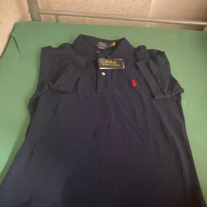 Marinblå pikétröja Polo Ralph Lauren - Snygg marinblå pikétröja från Polo Ralph Lauren i bomull. Klassisk krage, två knappar och röd broderad logga på bröstet. Modellen är custom slim fit och har korta ärmar. Perfekt för en clean och stilren look.