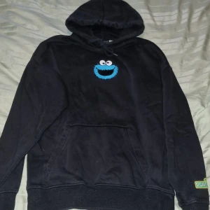 Svart Cookie Monster hoodie H&M XS - Svart hoodie från H&M x Sesame Street i relaxed fit. Broderad Cookie Monster på bröstet och patch med Sesame Street-logga på ärmen. Klassisk känguruficka, huva med snörning och mjukt bomullsmaterial. Perfekt för dig som gillar streetstyle och nostalgi.