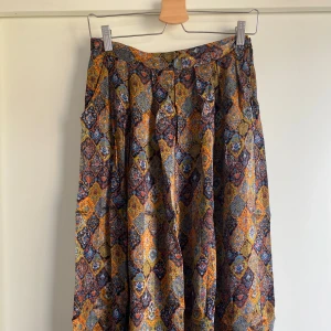 Färgglad långkjol med orientaliska mönster - Säljer en långkjol med färgstarkt mönster i blått, orange, gult och svart. Kjolen har en lös passform och är tillverkad i ett lätt tyg som faller snyggt. Perfekt för dig som gillar bohemiska vibes och vill sticka ut med coola färger och mönster. Har en lätt defekt i midjan men är fullt användbar!