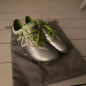 Silver fotbollsskor från New Balance Elite - Snygga fotbollsskor från New Balance i metallic silver med limegröna detaljer och snörning. Skorna har en slimmad form, platt sula med gröna dobbar och coolt mönstrad kromad häl. Perfekt för dig som vill sticka ut på planen.