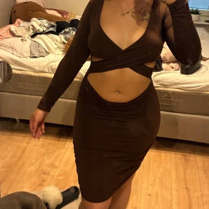 Brun omlottklänning med cut-outs - Säljer en brun kortklänning med långärmad design och snygga cut-outs vid midjan. Klänningen har omlott framtill med v-ringad hals och tight passform som framhäver figuren. Perfekt för dig som gillar trendiga och vågade plagg. Den är som en filt på insidan så passar även när det är lite kyligare ute. 