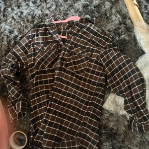Brun rutig skjorta med fickor XS-S - Snygg brun skjorta med vitt rutmönster och två bröstfickor med knappar. Skjortan har lång ärm och klassisk krage, perfekt för dig som gillar en chill och avslappnad stil. Materialet känns mjukt och bekvämt mot huden.